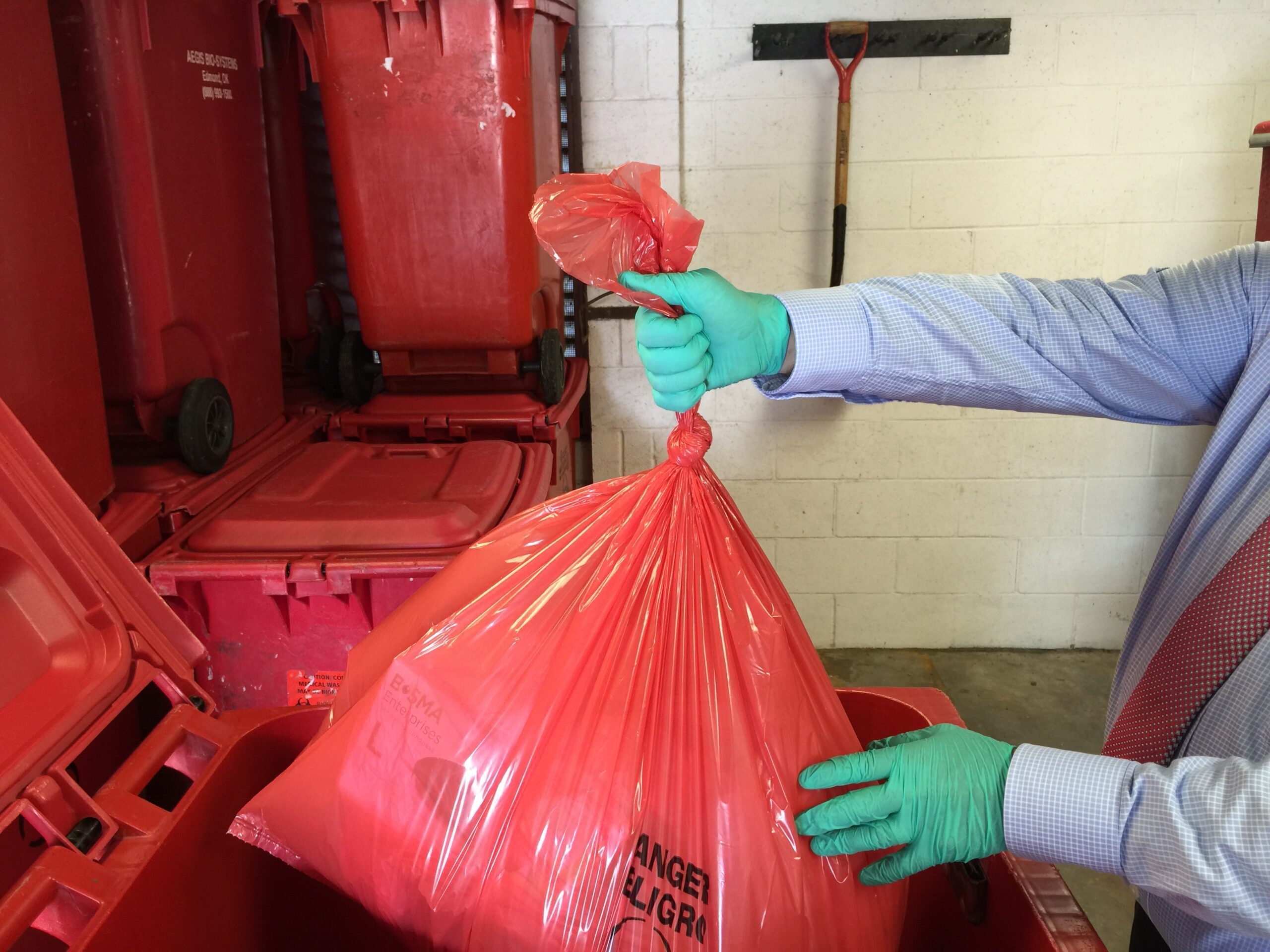 Gloved worker secures red bag for disposal into medical waste containers and packaging bins.