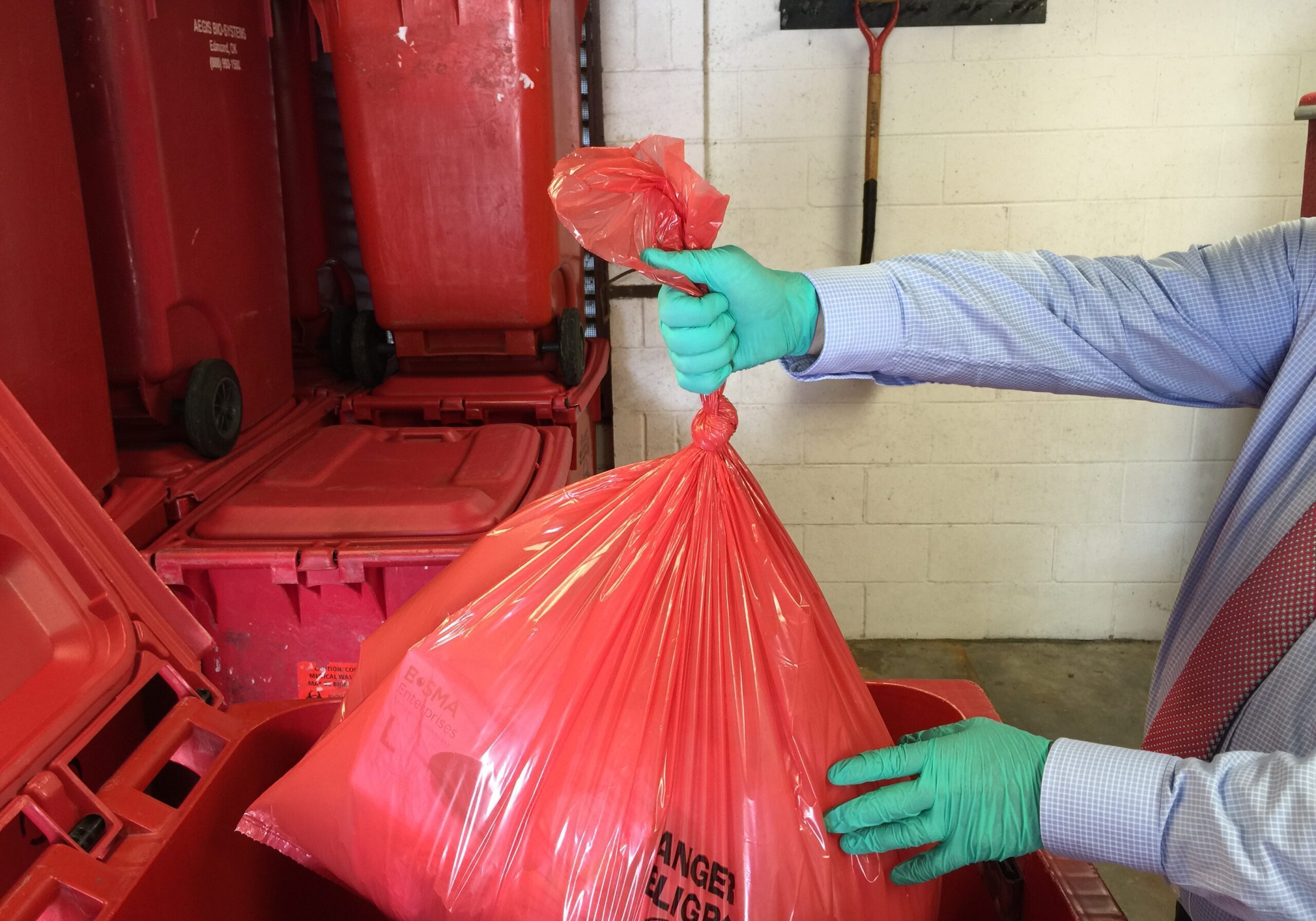 Gloved worker secures red bag for disposal into medical waste containers and packaging bins.